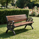 A wooden park bench with ornate black metal arms and legs, set on a green lawn in front of a trimmed hedge and garden flowers.