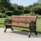 A wooden park bench with ornate black metal arms and legs, positioned on a light concrete path with a lush green garden in the background.