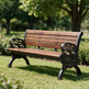 A wooden garden bench with ornate black cast iron legs and armrests, set on a green grassy lawn in a park with trees in the background.