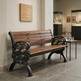 A wooden and cast iron park bench with ornate scrollwork arms and legs, shown in a museum or gallery setting next to a small side table with a pitcher and glasses.