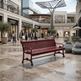 A red slatted outdoor bench sits in a modern shopping mall atrium with stone tile floors, potted trees, and shoppers walking in the background.