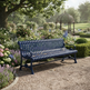 A dark blue metal garden bench with a slatted back and seat, positioned on a gravel path in a lush, sunlit garden with flowers, a tree, and a gazebo in the background.