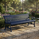 A dark navy blue metal park bench with a slatted back and seat, positioned on a stone pathway in a lush, sunlit garden with flowers and trees in the background.