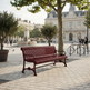 A red metal park bench in a paved public square with trees, a fountain, historic buildings, and a few people in the background.