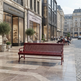 A red slatted outdoor bench sits on a paved city plaza in front of elegant storefronts, with seated people and urban buildings in the background.