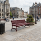 A red slatted park bench sits on a cobblestone plaza in a historic city square, with pedestrians, buildings, a lamp post, and a planter visible in the background.