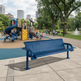 A blue metal park bench in the foreground of a city playground scene, with children playing on a slide and climbing structure in the background among trees and a fountain.