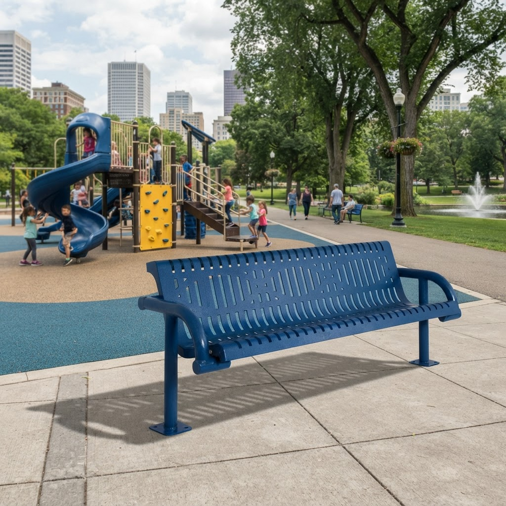 A blue metal park bench in the foreground of a city playground scene, with children playing on a slide and climbing structure in the background among trees and a fountain.