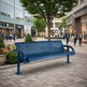 A blue metal park bench with a slatted back and seat, placed on a paved plaza in a city shopping district with pedestrians, trees, and storefronts in the background.