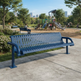 A blue metal park bench with a slatted back and seat sits on a concrete path in a city park, with a playground, trees, and distant buildings in the background.
