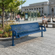 A blue metal park bench in an outdoor urban plaza, with a seated man on a bench nearby and a large stone building in the background.
