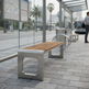 A modern outdoor bench with a light brown wooden slatted seat and brushed silver metal frame sits on a paved transit station platform. A bus is visible in the blurred background, along with a glass wall, maps, and a person standing at the right edge.
