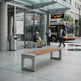 A modern outdoor transit station scene with a long bench in the foreground, glass shelter, seated and walking passengers, and a city bus at a bus stop.