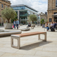 A modern outdoor bench with a warm brown wooden seat and brushed silver metal frame, situated in a paved public square with people and buildings in the background.