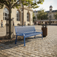 A blue metal park bench sits on a cobblestone plaza beside a tree and a brown trash can, with a city square and historic buildings in the background.