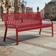 A modern red metal bench with a slatted back and seat, positioned on a cobblestone street in an urban European setting with historic buildings and a tram in the background.