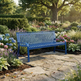 A blue metal garden bench with slatted back and seat, placed on a stone path in a lush, sunlit garden with flowers, shrubs, and trees.