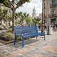A blue slatted metal park bench in a paved urban plaza with trees, buildings, pedestrians, and a trash can in the background.
