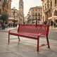 A bright red slatted metal bench sits on a stone plaza in a historic European town square, with buildings, street lamps, and walking pedestrians in the background.