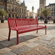 A bright red slatted metal bench in a European town square, with historic stone buildings, pedestrians, and trees in the background.