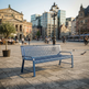 A blue metal park bench in a spacious city square, with historic buildings, pedestrians, cyclists, and a potted tree in the background.
