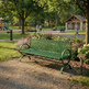 A green metal park bench sits on a gravel path in a sunny, tree-lined public park with flower beds, a small pavilion, and distant people walking and playing.