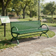 A green metal park bench sits on a gravel path in a sunny park with trees, grass, and a pond in the background. A small signpost is visible to the left.