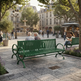 A green slatted park bench in a paved public square, with people sitting and walking around in a real urban environment. Trees, buildings, a fountain, and a kiosk are visible in the background.