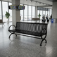 A black metal airport bench with a slatted back and seat, positioned in a spacious, modern airport terminal with large windows, flight information boards, and travelers in the background.