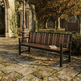 A wooden park bench with a dark metal frame sits on a stone pathway in an outdoor garden setting with autumn trees and a stone wall in the background.