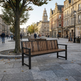 A modern wooden slat bench with a black metal frame sits on a stone plaza in a city setting, with people sitting and walking in the background near historic buildings and a tree.