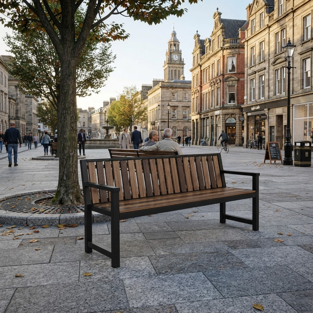 A modern wooden slat bench with a black metal frame sits on a stone plaza in a city setting, with people sitting and walking in the background near historic buildings and a tree.