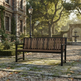 A dark brown outdoor wooden bench with a slatted back and seat, positioned on a stone path in a garden courtyard with a historic stone building, trees, and a lamp post in the background.