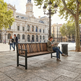 A wooden slat bench with a black metal frame sits in a public square, with a woman sitting on it and reading a newspaper. A large historic building, trees, and street lamps are visible in the background.