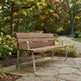 A wooden outdoor bench with a weathered brown finish and a silver metal frame, situated on a stone path in a park with trees and shrubs in the background.