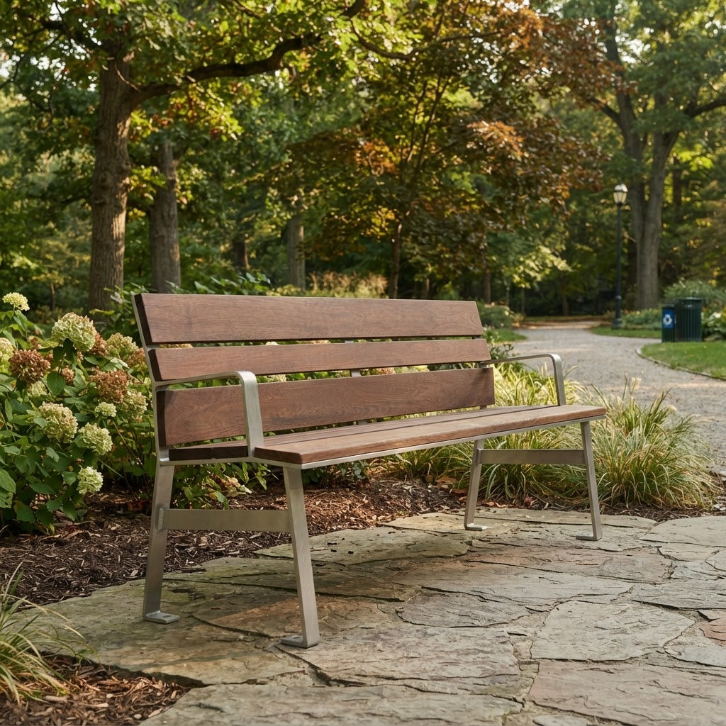 A wooden outdoor bench with a weathered brown finish and a silver metal frame, situated on a stone path in a park with trees and shrubs in the background.