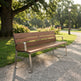 A modern wooden park bench with a brown wood-grain slatted back and seat, supported by a silver metal frame, situated on a gravel path in a sunny park with green grass and trees.