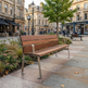 A modern wooden park bench with a brown slatted seat and backrest, supported by silver metal legs, situated on a paved city sidewalk in an urban plaza with buildings, a tree, and storefronts in the background.