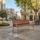 A modern wooden park bench with a brushed silver metal frame, situated in a sunny, historic European town square with stone paving, trees, and buildings in the background.