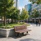 A brown wooden park bench with black metal legs sits beside a stone planter in a city plaza, with trees, flowers, pedestrians, and modern glass buildings in the background.