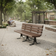A brown slatted outdoor bench with black metal legs sits on a paved walkway in a park-like setting with trees, a small shelter, and a couple seated in the background.