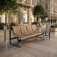 A modern outdoor bench with a curved dark metal frame and light brown wooden slats, situated on a stone plaza in an urban setting with buildings, trees, bollards, and a small shopping bag beside it.