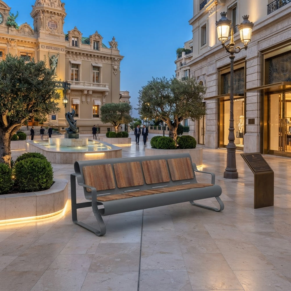 A modern outdoor bench with a gray metal frame and brown wooden slats, situated in a beautifully lit urban plaza with historic buildings, a fountain, and trees at dusk.