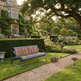 A modern outdoor bench with a dark gray metal frame and warm brown wooden slats, placed on a gravel path in a beautifully landscaped garden with a large stone mansion in the background.