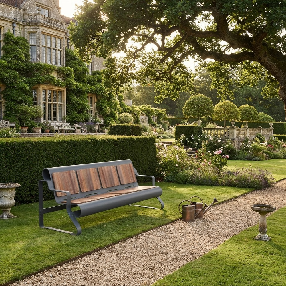 A modern outdoor bench with a dark gray metal frame and warm brown wooden slats, placed on a gravel path in a beautifully landscaped garden with a large stone mansion in the background.