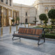 A modern outdoor bench with a dark gray metal frame and brown wood-look seat and back panels, placed in a grand public plaza with a fountain, historic building, and manicured gardens in the background.