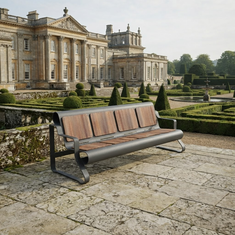 A modern outdoor bench with a dark gray metal frame and brown wooden slats, positioned on a stone patio in front of a grand classical estate with manicured gardens.