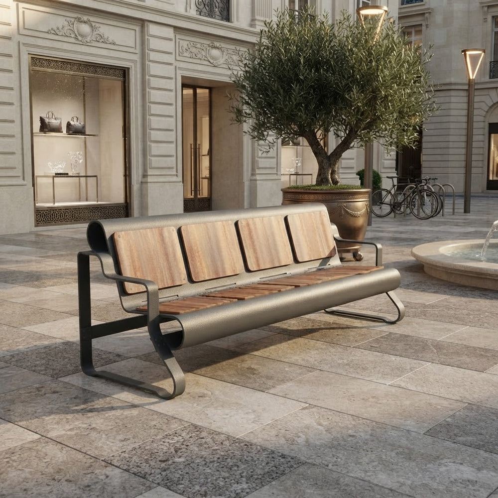 A modern outdoor bench with a curved metal frame and light wood slats, placed on a stone plaza in front of a building with storefront windows and a large potted tree.