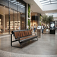 A modern outdoor-style bench with a curved black metal frame and brown wooden slats sits in a spacious, upscale shopping mall plaza. The background features a large glass-front boutique, shoppers walking under a glass roof, and green trees.