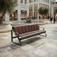 A modern outdoor bench with dark metal framing and brown wood-look seat and back panels, placed in a upscale plaza with stone tiles, trees, a fountain, and buildings in the background.